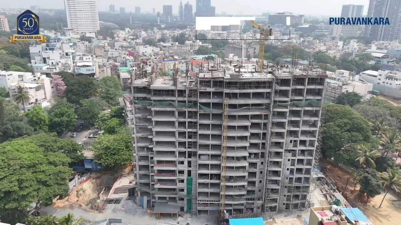 Front aerial view of the Purva Orient Grand tower under construction on Lal Bagh Main Road, its concrete frame and scaffolding rising above the trees of Sudhama Nagar, from a Puravankara status update